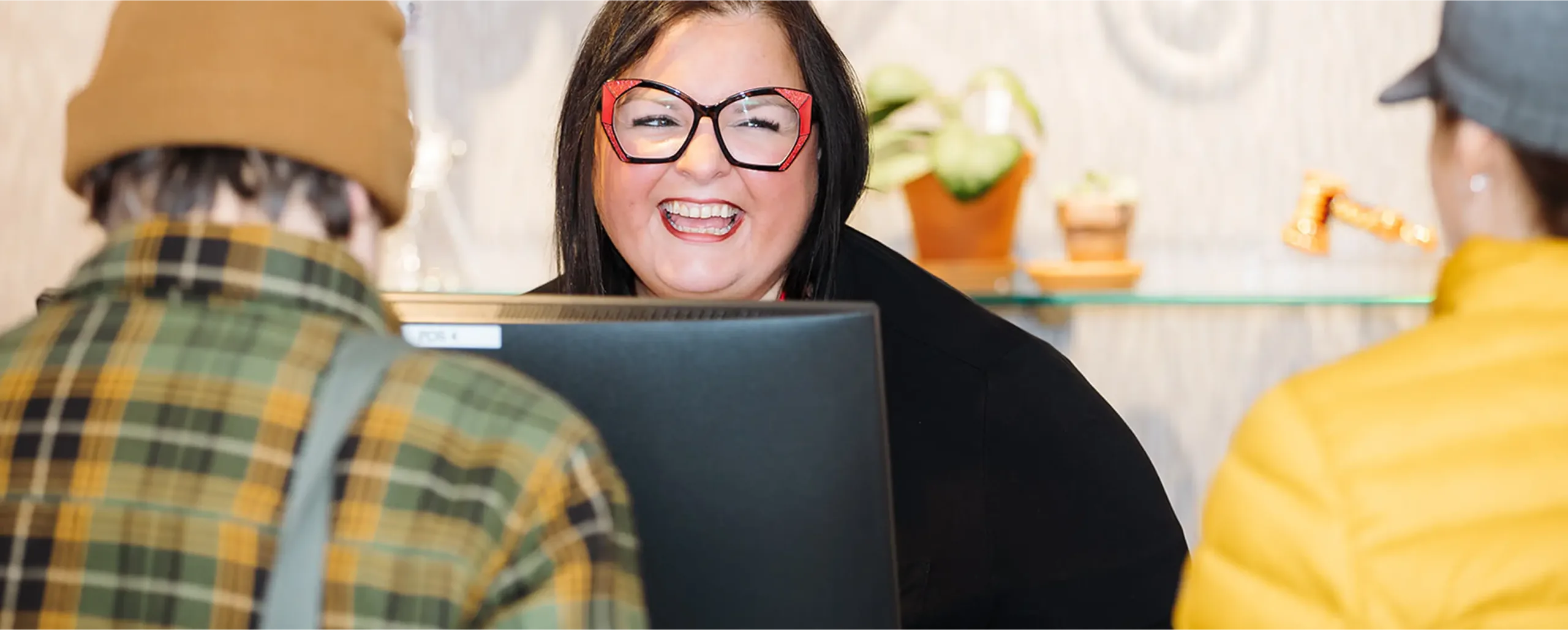 A smiling woman with red glasses stands behind a counter, engaging with two people wearing jackets and hats.