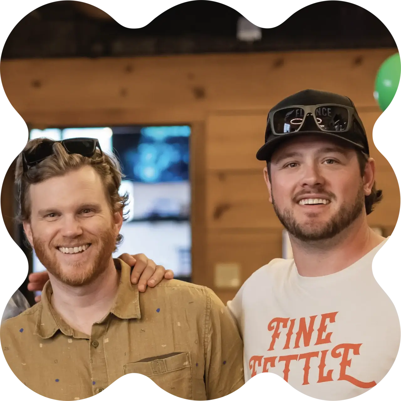 Two smiling men pose indoors; one wears a tan shirt, the other a “FINE FETTLE” T-shirt—showing camaraderie at a cannabis dispensary. One has an arm around the other.