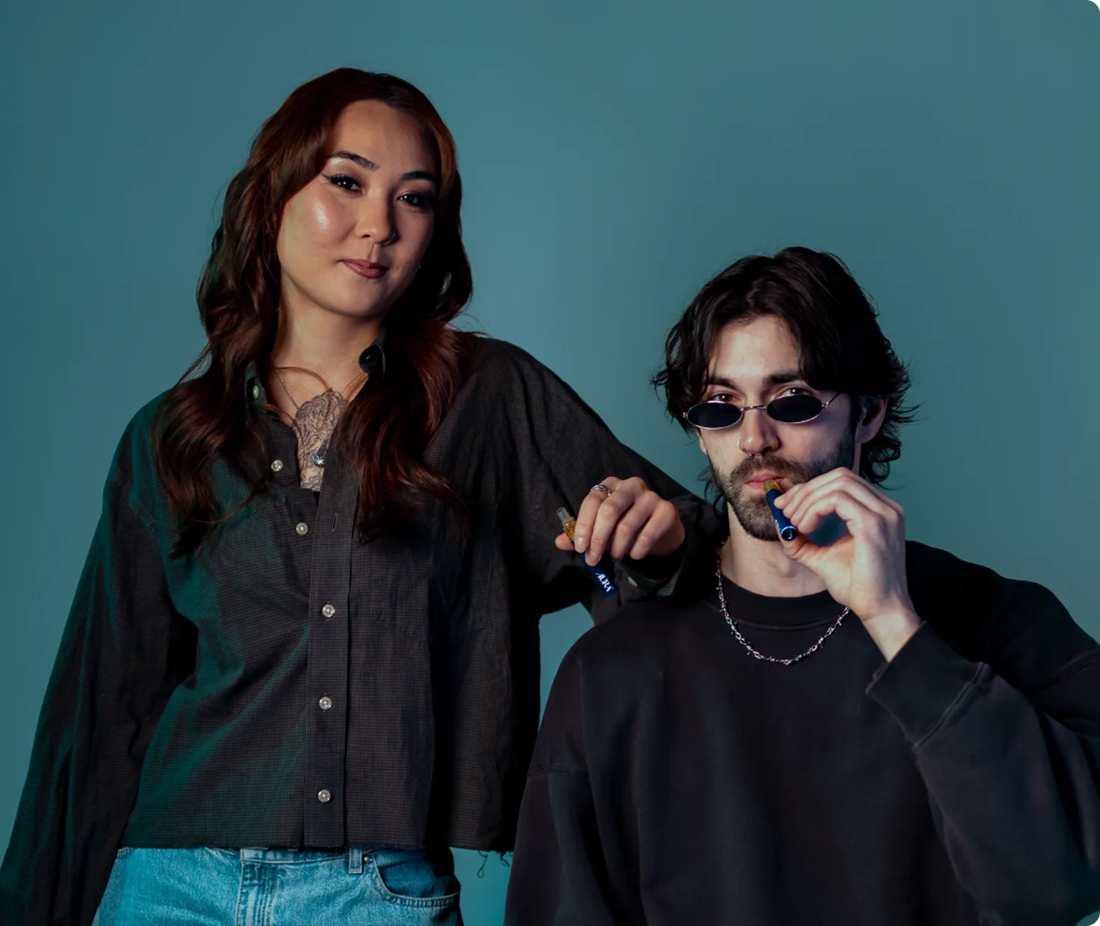 Two people pose against a blue background, one standing with a vape pen and the other sitting, wearing sunglasses and using a vape pen.