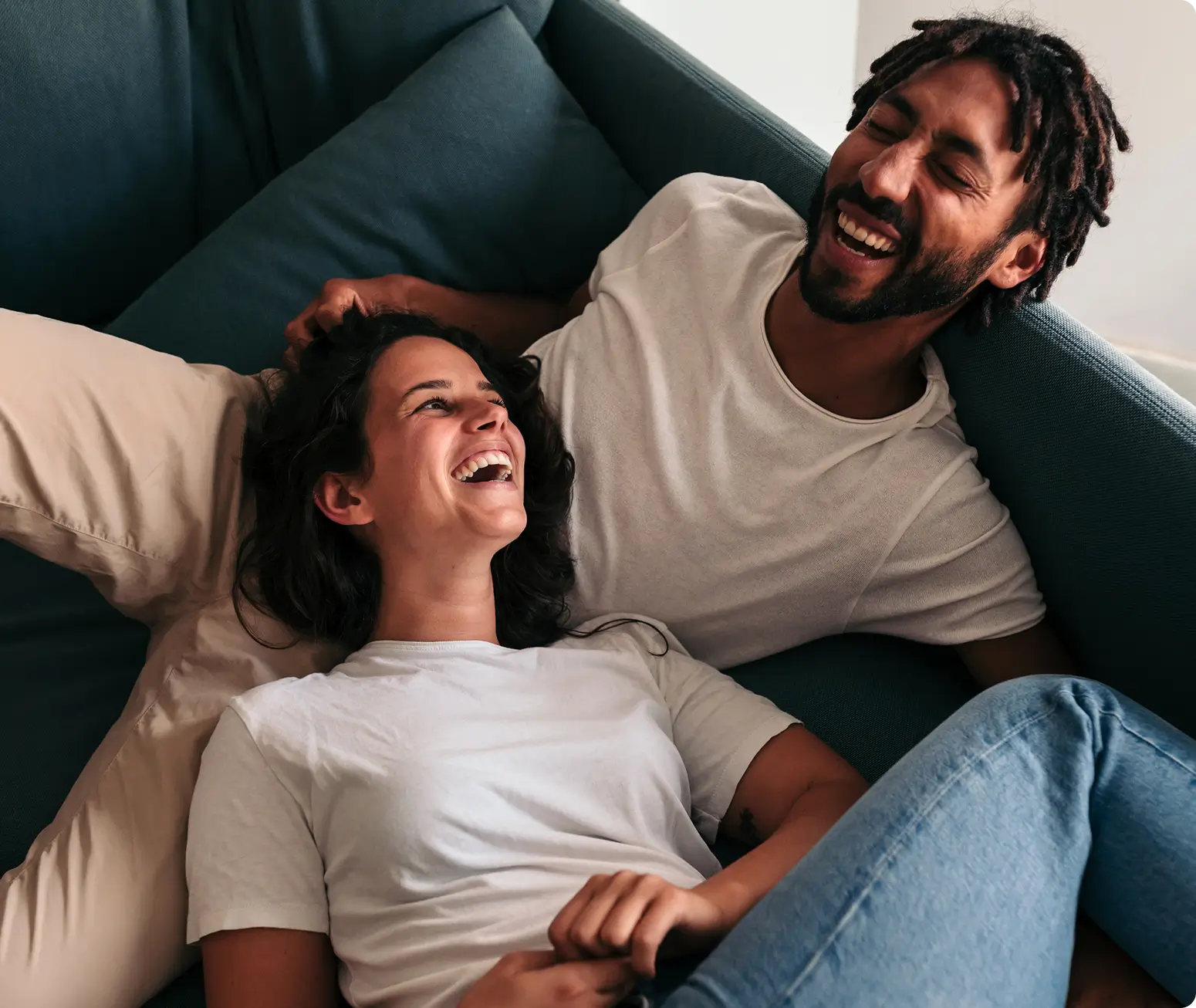A smiling couple lounges on a sofa, laughing together while relaxing in casual clothes.