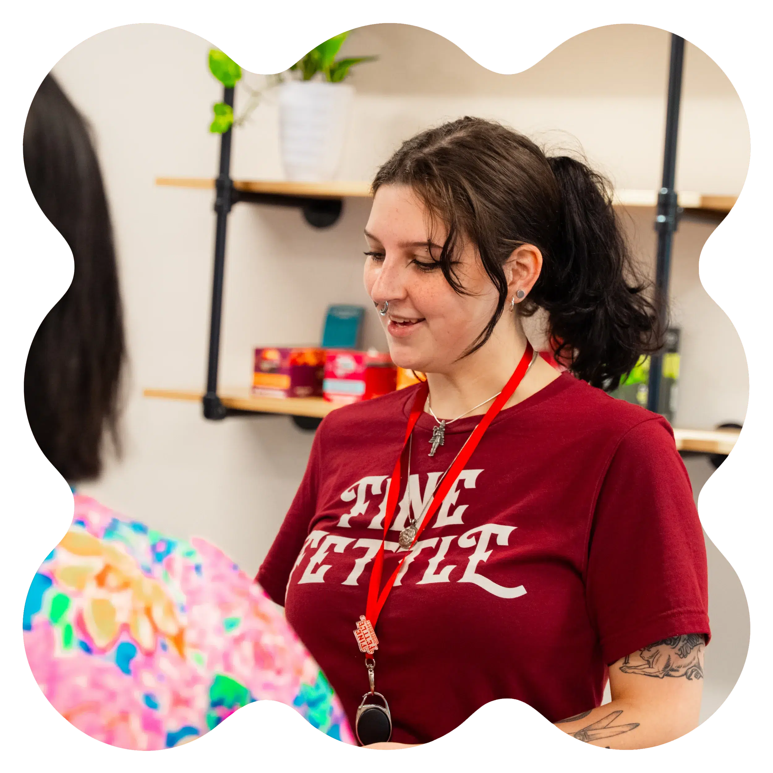A person wearing a red shirt with white text and a red lanyard is smiling and talking to another person in a colorful shirt indoors.