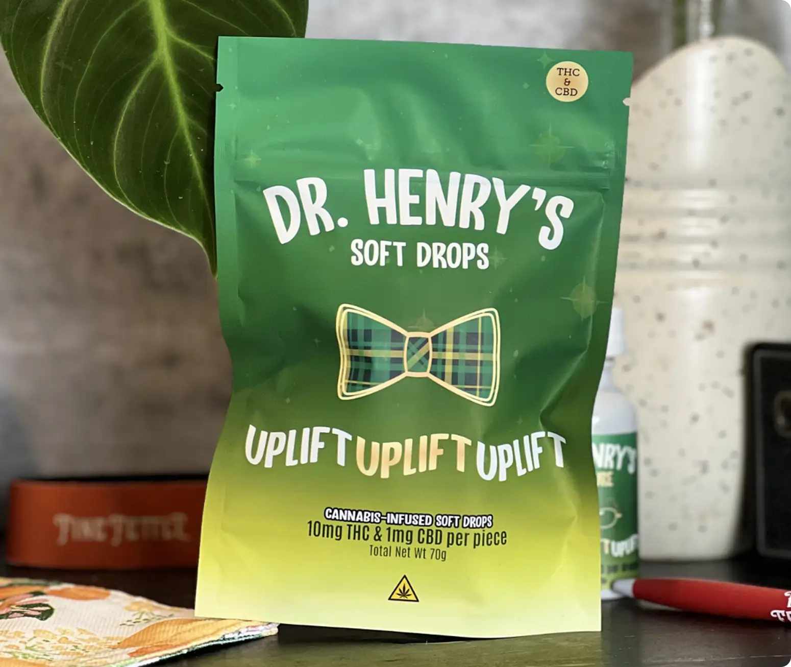 A green bag of Dr. Henry’s Soft Drops featuring a plaid bow tie and “Uplift” repeated, with 10mg THC and 2mg CBD per piece stated on the front. The bag is on a table with other items nearby.