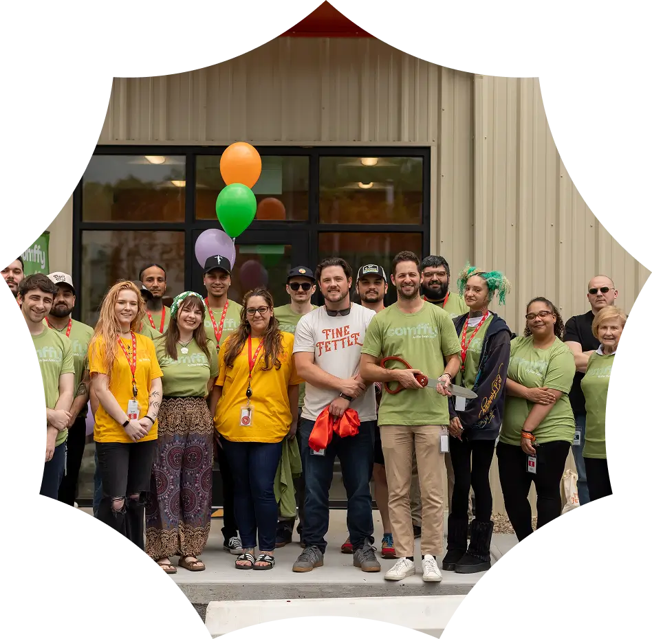 A group of people smiling and posing together outside a building, some wearing matching shirts, with balloons in the background.
