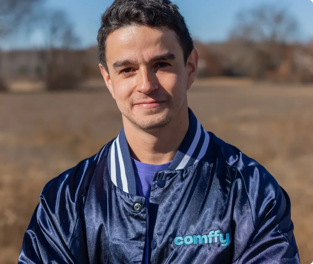 A young man wearing a shiny blue comify jacket stands outdoors in a field, smiling slightly at the camera on a sunny day, embodying the relaxed vibe of top cannabis brands.